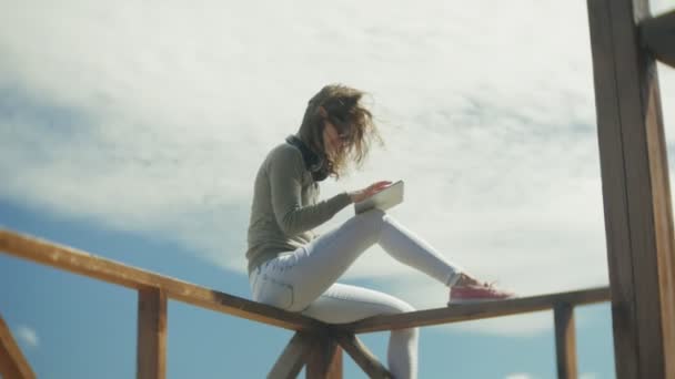 Joven hermosa mujer se sienta en la playa en los auriculares y utiliza una tableta — Vídeos de Stock