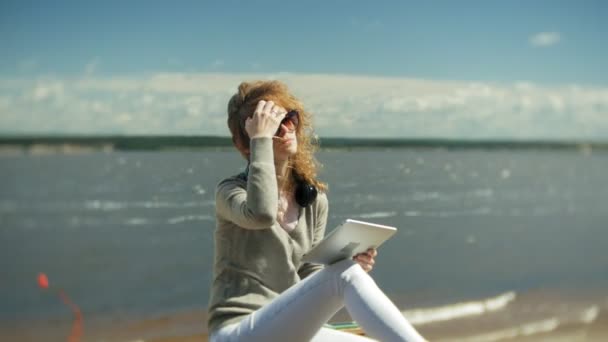 Genç güzel kadın kulaklık, sahilinde oturur ve bir tablet kullanır — Stok video
