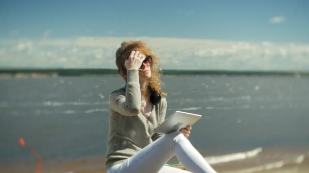 Young beautiful woman sits on the beach in headphones and uses a tablet — Stock Video