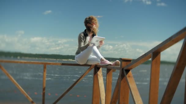 Jovem mulher bonita senta-se na praia em fones de ouvido e usa um tablet — Vídeo de Stock