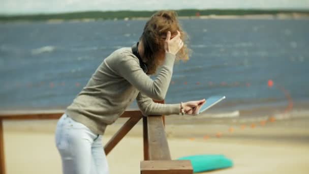 Jovem mulher bonita senta-se na praia em fones de ouvido e usa um tablet — Vídeo de Stock