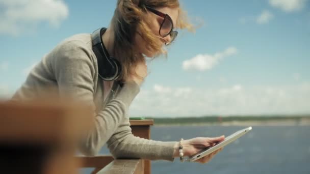 Giovane bella donna si siede sulla spiaggia in cuffia e utilizza un tablet — Video Stock