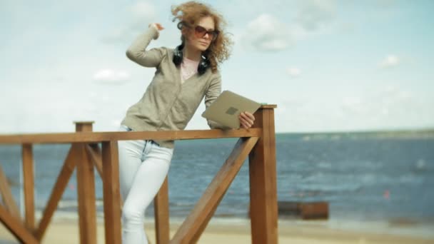 Young beautiful woman sits on the beach in headphones and uses a tablet — Stock Video