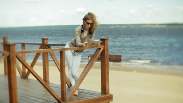Jovem mulher bonita senta-se na praia em fones de ouvido e usa um tablet — Vídeo de Stock
