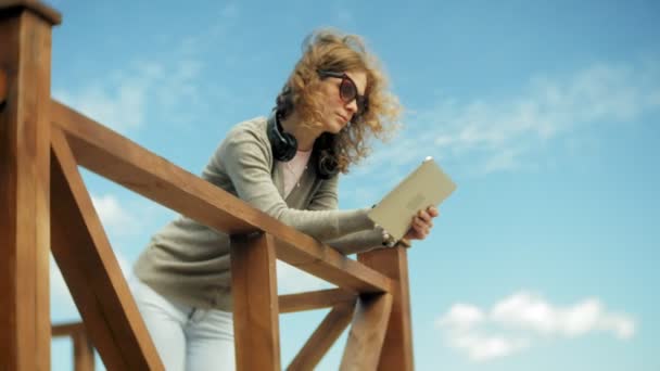 Genç güzel kadın kulaklık, sahilinde oturur ve bir tablet kullanır — Stok video