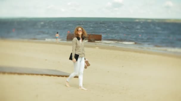 Молодая красивая женщина гуляет по пляжу в наушниках и использует планшет — стоковое видео