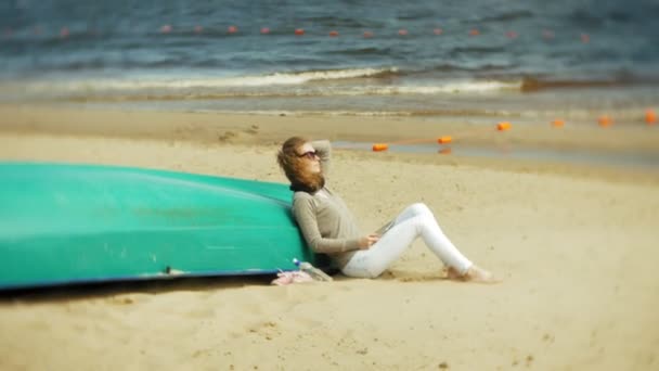 Νεαρή όμορφη γυναίκα κάθεται στην παραλία ακουστικά κοντά στο σκάφος και χρησιμοποιεί ένα δισκίο — Αρχείο Βίντεο