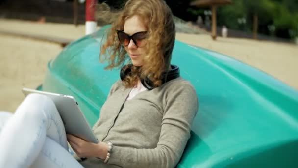 Młoda piękna kobieta siedzi na plaży w słuchawki w pobliżu łodzi i korzysta z tabletu — Wideo stockowe