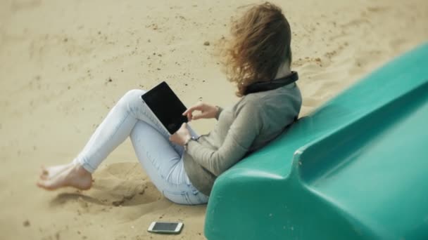 Genç güzel kadın kulaklık tekne yakınındaki kumsalda oturur ve bir tablet kullanır — Stok video