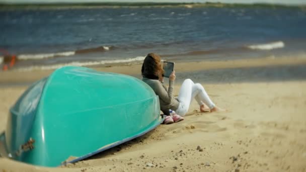 Νεαρή όμορφη γυναίκα κάθεται στην παραλία ακουστικά κοντά στο σκάφος και χρησιμοποιεί ένα δισκίο — Αρχείο Βίντεο