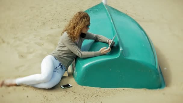 Giovane bella donna si siede sulla spiaggia in cuffie vicino alla barca e utilizza un tablet — Video Stock