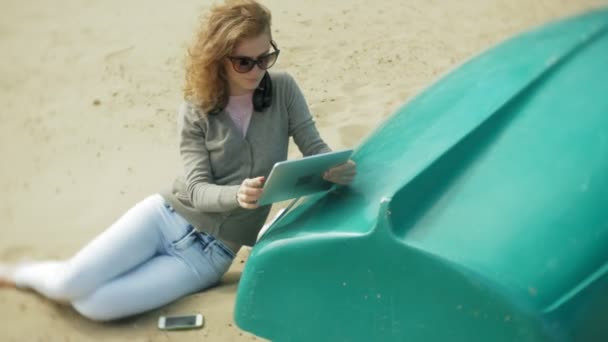 Jovem mulher bonita senta-se na praia em fones de ouvido perto do barco e usa um tablet — Vídeo de Stock