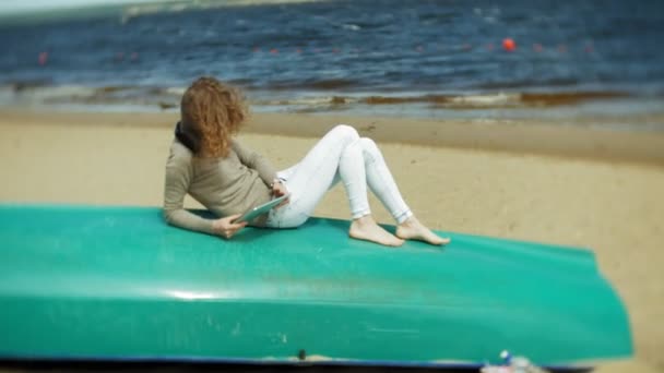 Ung vacker kvinna sitter på stranden i hörlurar nära båten och använder en surfplatta — Stockvideo