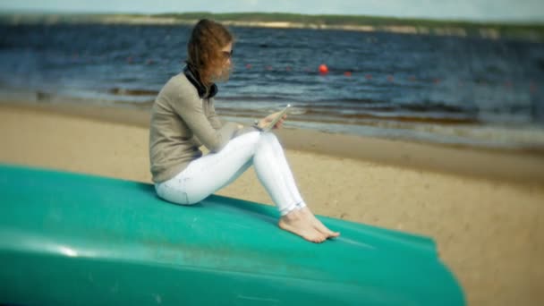 Молодая красивая женщина сидит на пляже в наушниках возле лодки и использует планшет — стоковое видео