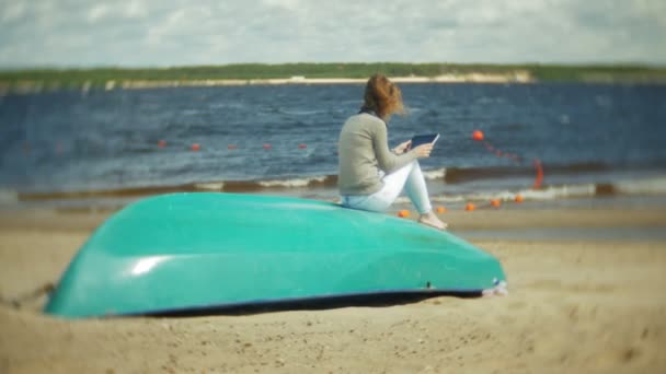 Giovane bella donna si siede sulla spiaggia in cuffie vicino alla barca e utilizza un tablet — Video Stock