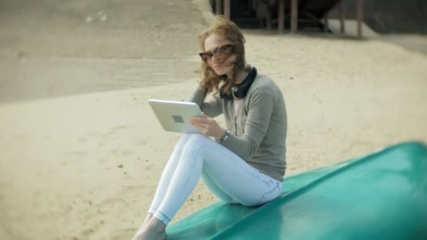 Mooie jonge vrouw zit op het strand in de hoofdtelefoon in de buurt van de boot en maakt gebruik van een tablet — Stockvideo