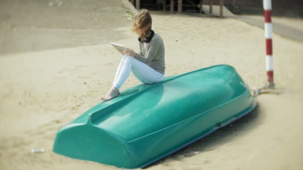 Joven hermosa mujer se sienta en la playa en auriculares cerca del barco y utiliza una tableta — Vídeo de stock