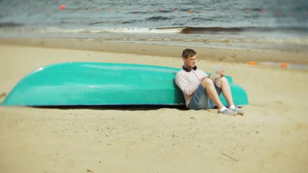 Viejo se sienta en la playa en auriculares cerca del barco y utiliza una tableta — Vídeos de Stock