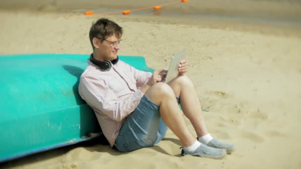 老人坐在海滩上的耳机附近的船和使用平板电脑4K — 图库视频影像