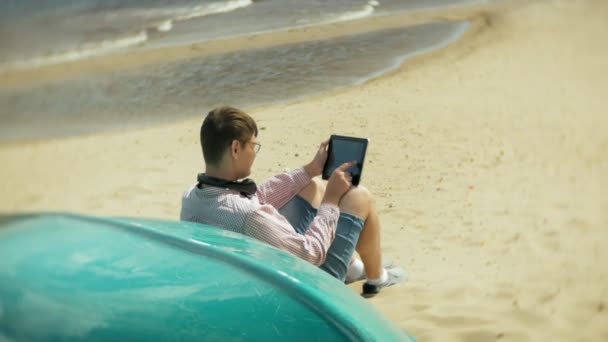 Gubben sitter på stranden i hörlurar nära båten och använder en surfplatta — Stockvideo