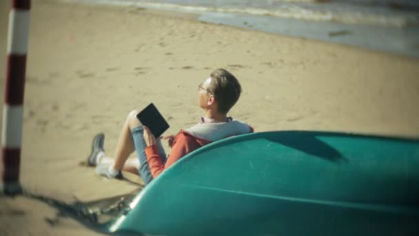 Stary człowiek siedzi na plaży w słuchawki w pobliżu łodzi i korzysta z tabletu — Wideo stockowe