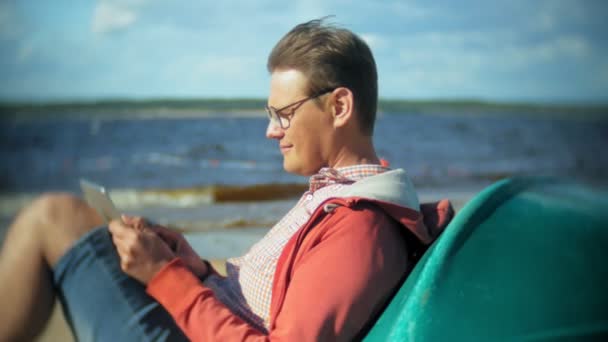 Viejo se sienta en la playa en auriculares cerca del barco y utiliza una tableta — Vídeos de Stock