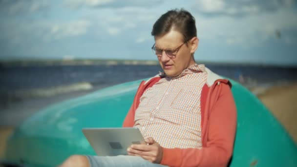 Stary człowiek siedzi na plaży w słuchawki w pobliżu łodzi i korzysta z tabletu — Wideo stockowe