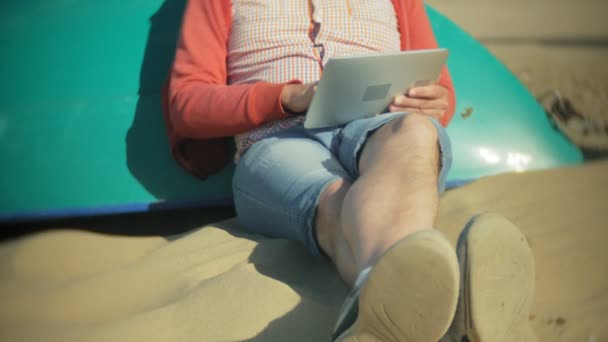 Старик сидит на пляже в наушниках возле лодки и пользуется планшетом — стоковое видео