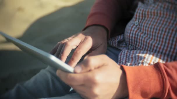 Vecchio si siede sulla spiaggia in cuffie vicino alla barca e utilizza un tablet — Video Stock