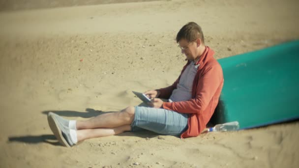 Старик сидит на пляже в наушниках возле лодки и пользуется планшетом — стоковое видео