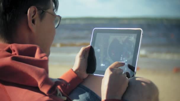 Velho senta-se na praia em fones de ouvido perto do barco e usa um tablet — Vídeo de Stock