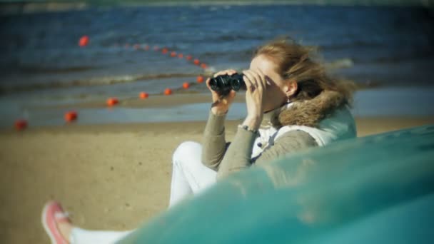 Jonge vrouw zit op het strand buiten de boot en kijkt door een verrekijker — Stockvideo