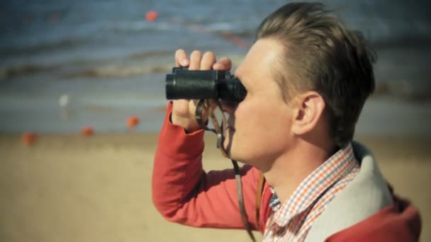 Літній чоловік сидить на пляжі біля човна і дивиться через бінокль — стокове відео
