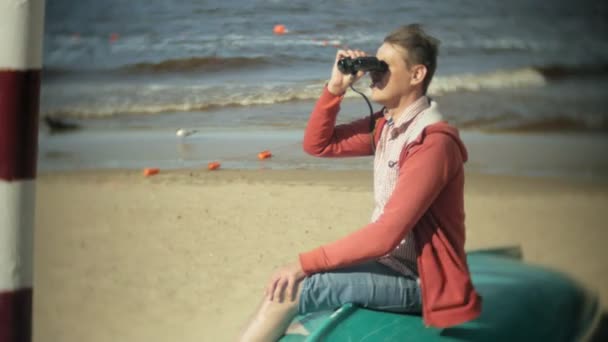 Starszy pan siedzi na plaży na obrzeżach łodzi i patrzy przez lornetkę — Wideo stockowe