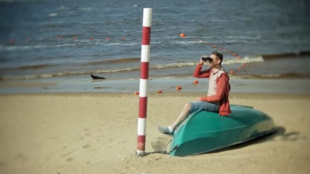 Een oudere man zit op het strand buiten de boot en ziet er door middel van verrekijkers — Stockvideo