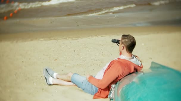 En äldre man sitter på stranden utanför båten och ser genom kikare — Stockvideo