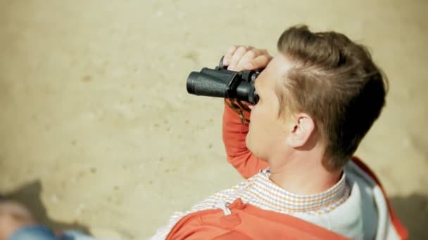 An elderly man sits on the beach outside the boat and looks through binoculars — Stock Video