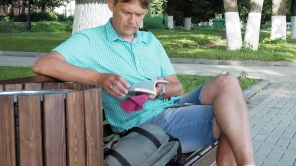 Gözlüklü bir adam parkta bir bankta oturur ve bir kitap okur — Stok video