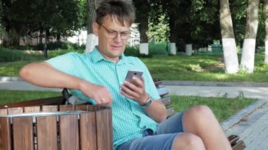 Gözlüklü bir adam parkta bir bankta oturur ve bir telefon kullanır