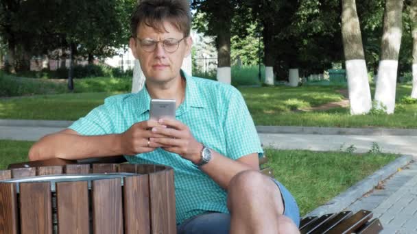 En man i glasögon sitter på en bänk i parken och använder en telefon — Stockvideo