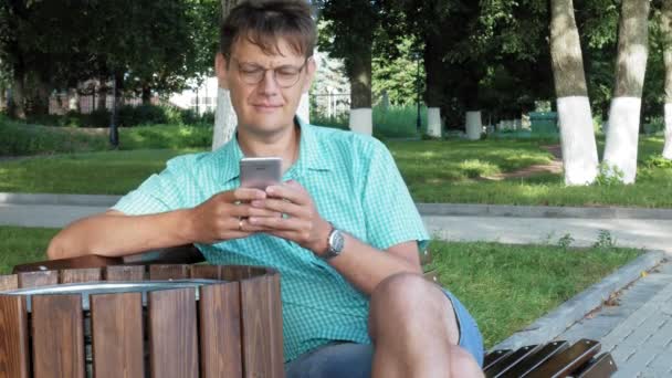 Ein Mann mit Brille sitzt auf einer Bank im Park und benutzt ein Handy — Stockvideo