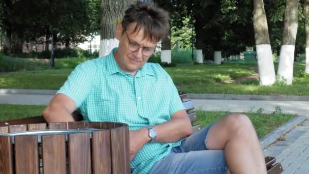 Un hombre con gafas se sienta en un banco en el parque y usa un teléfono — Vídeo de stock