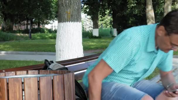 Un hombre con gafas se sienta en un banco en el parque y usa un teléfono — Vídeos de Stock