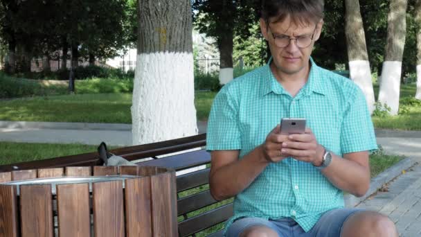 Een man in glazen zit op een bankje in het park en maakt gebruik van een telefoon — Stockvideo