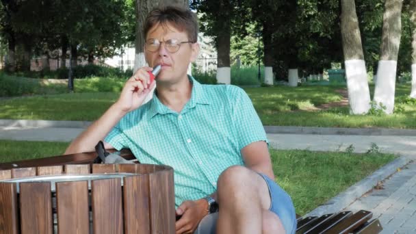 Un hombre con gafas se sienta en un banco en el parque y usa un teléfono — Vídeo de stock