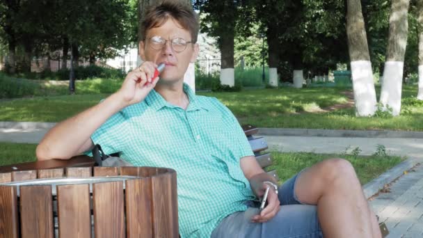 Чоловік в окулярах сидить на лавці в парку і використовує телефон — стокове відео