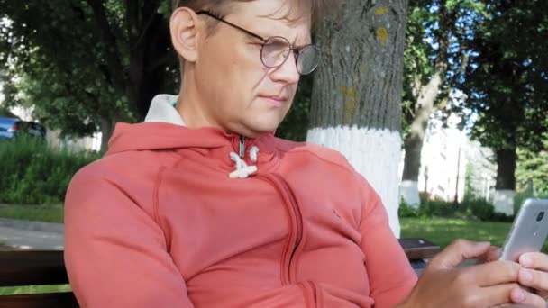 Seorang pria berkacamata duduk di bangku taman dan menggunakan telepon — Stok Video