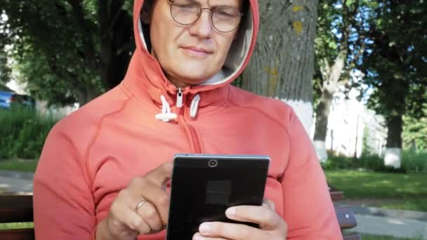 Un hombre con gafas se sienta en un banco en el parque y usa una tableta — Vídeos de Stock