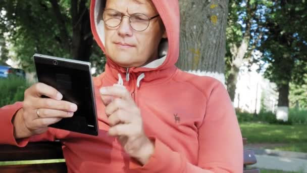 Un hombre con gafas se sienta en un banco en el parque y usa una tableta — Vídeos de Stock