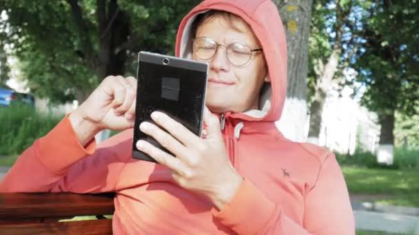 Um homem de óculos senta-se em um banco no parque e usa um tablet — Vídeo de Stock